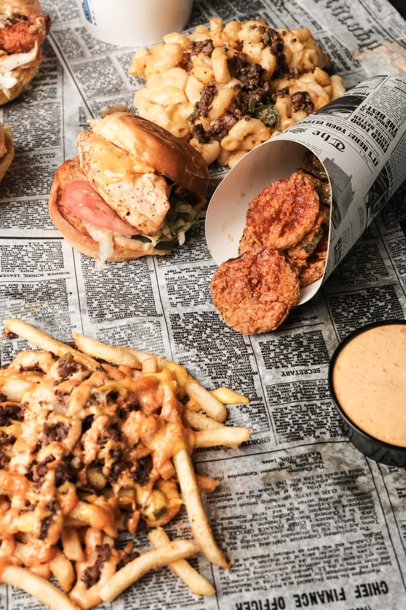 Assortment of burgers, loaded fries, mac and cheese, fried pickles, and dipping sauce on newspaper.