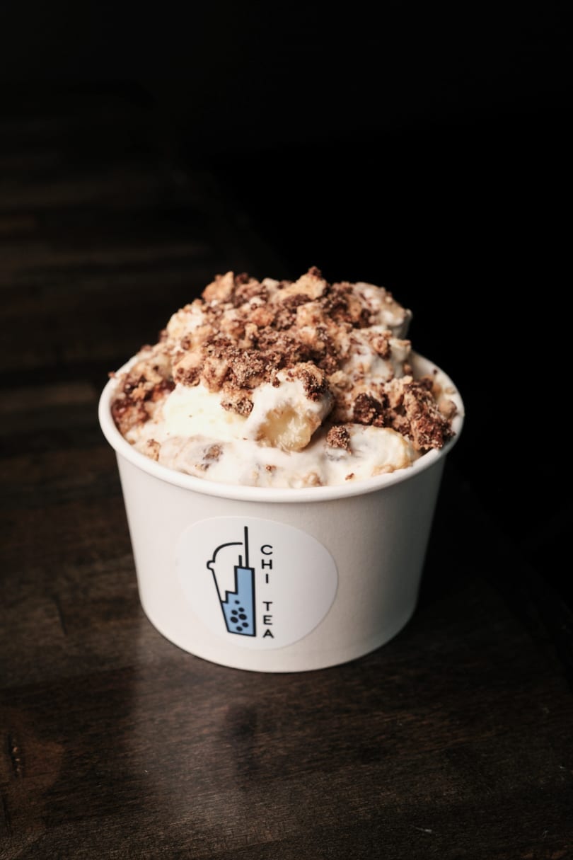 A white cup of Chi Tea banana pudding with cookie crumbles on a dark wooden surface.