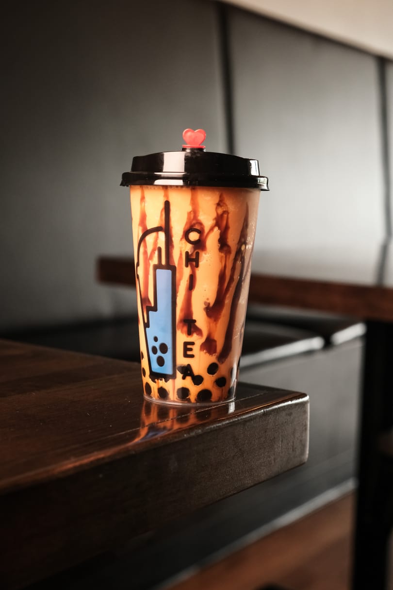 Boba tea with chocolate drizzle and a heart-shaped straw stopper sits on a dark wooden table.