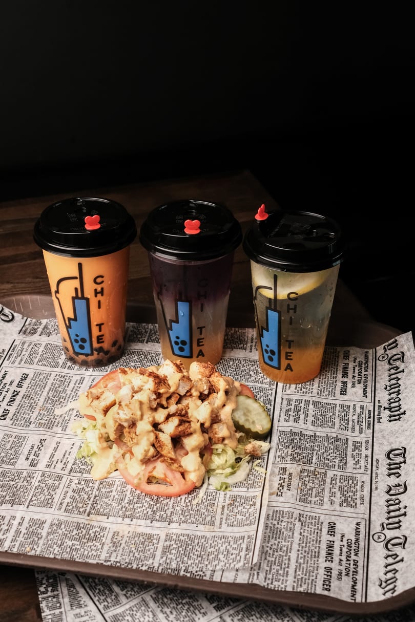 Three Chi Tea drinks and a chicken salad with tomatoes and pickles on newspaper-print paper.