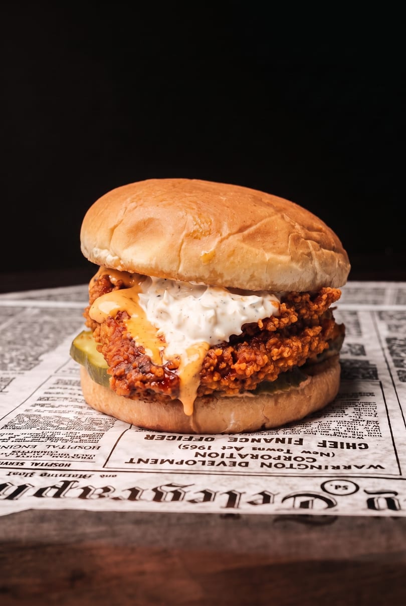 Fried chicken sandwich with pickles, white sauce, and orange sauce on a bun, served on newspaper.