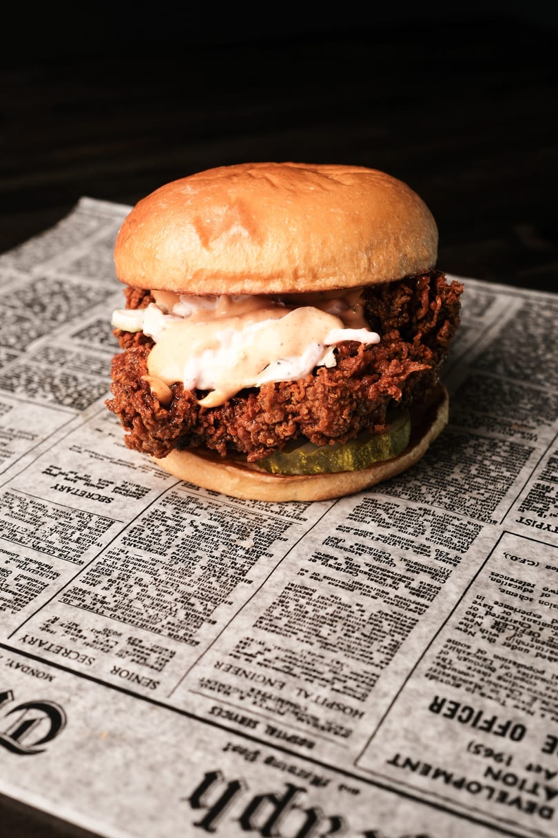 A crispy fried chicken sandwich with sauce and pickles rests on a newspaper.