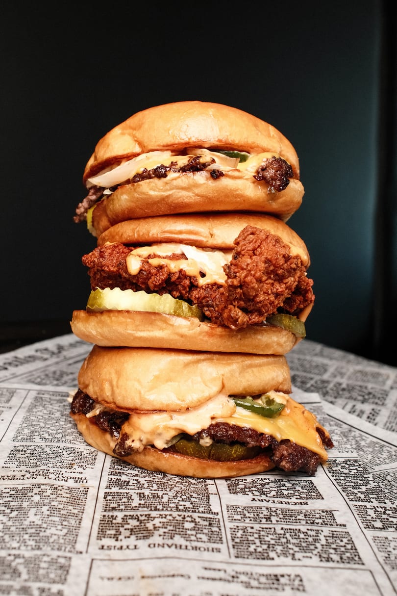 A towering stack of three burgers, including beef and fried chicken, on newspaper-patterned paper.