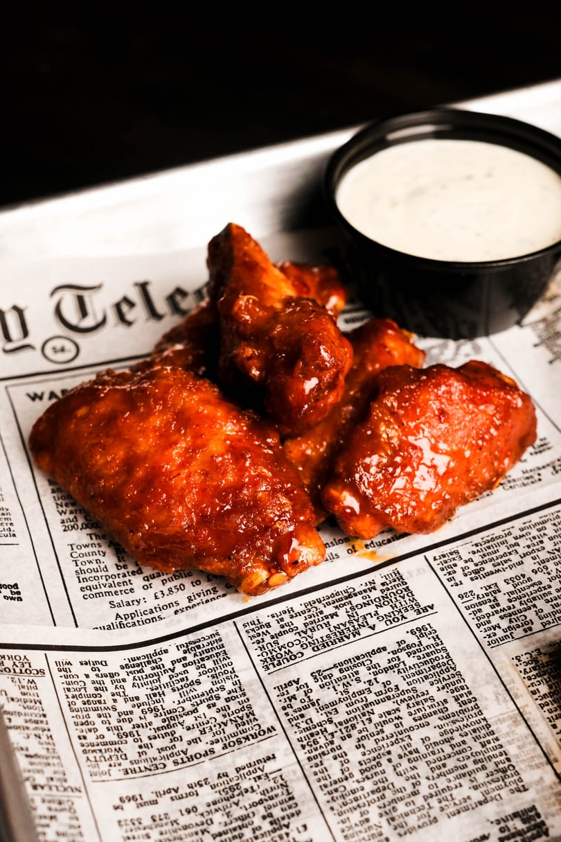 Glazed chicken wings and a side of ranch dressing served on newspaper-lined tray.