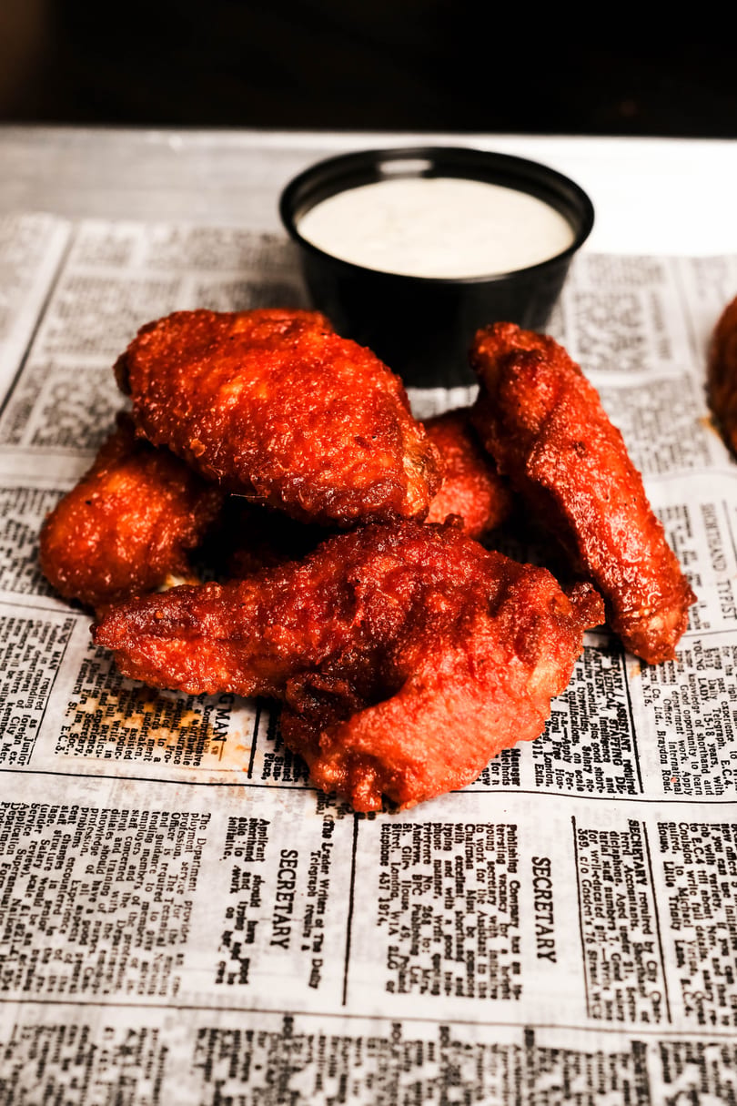 Spicy red chicken wings and a cup of ranch dressing on newspaper-lined tray.