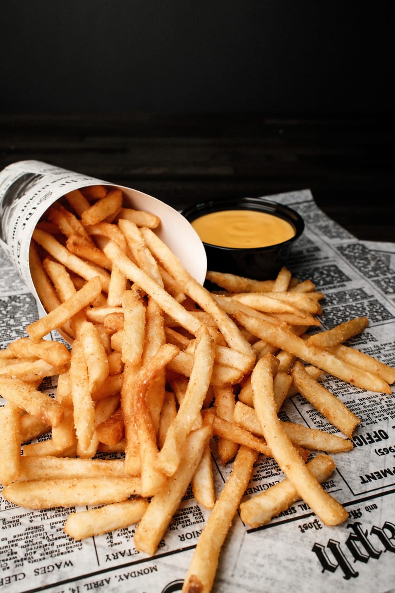 Crispy golden french fries spilling from a paper cone onto newspaper, with a side of cheese sauce.