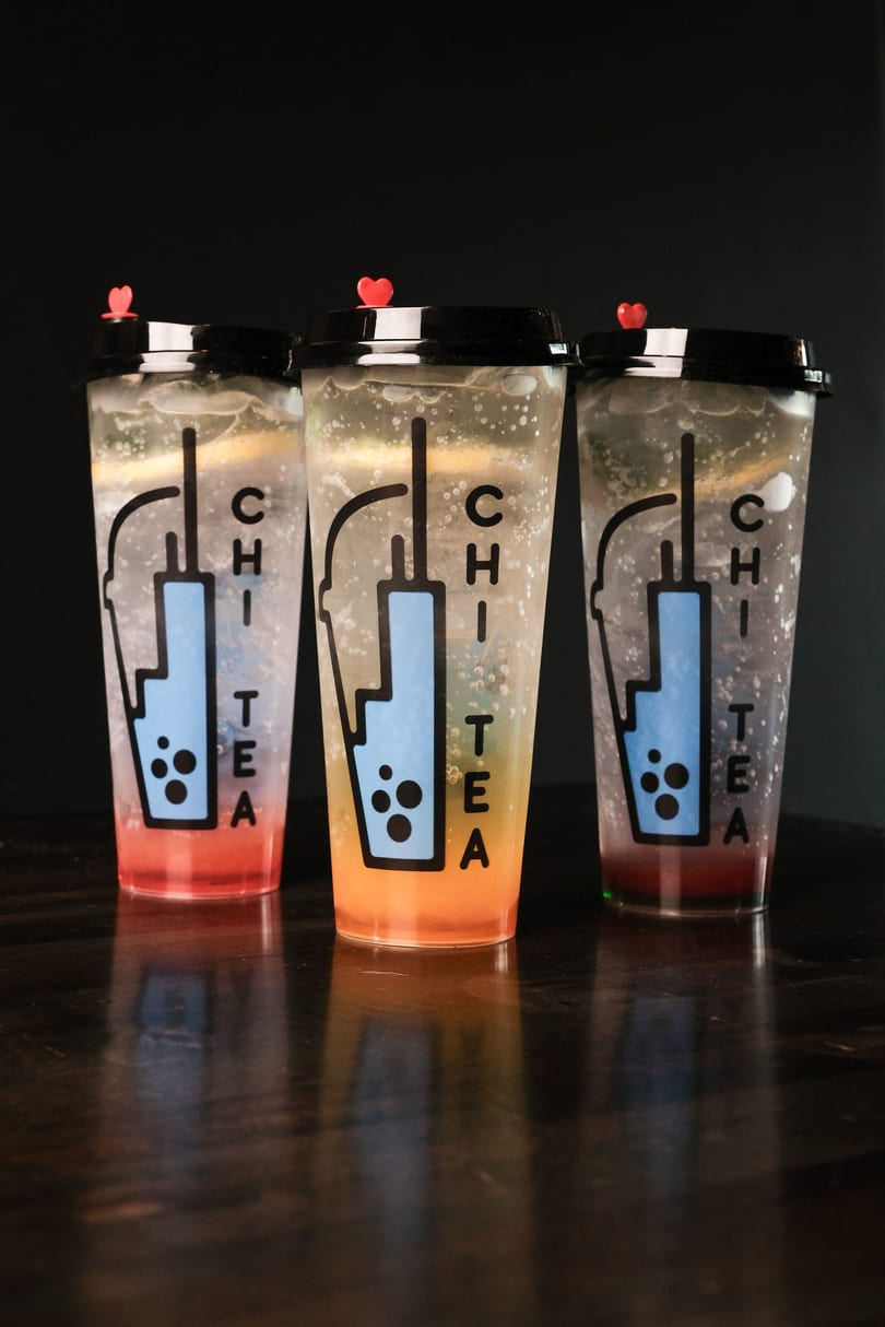 Three Chi Tea drinks in clear cups with red, orange, and purple liquid on a dark table.