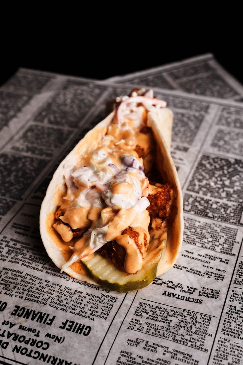 A fried chicken taco with coleslaw, sauce, and a pickle slice, on a newspaper.